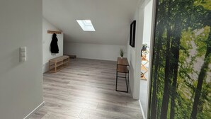 Una habitación amplia con techo inclinado, tragaluz, banco de madera, una mesita y una planta en la pared.