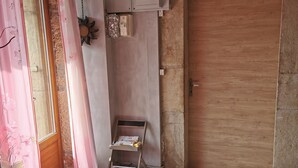 Una habitación con una puerta de madera, un escritorio pequeño de madera con libros y papeles, un cuadro eléctrico en la pared y una ventana con cortinas rosadas.