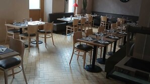 Een ruime eethoek met meerdere tafels en stoelen, grote ramen die natuurlijk licht binnenlaten, en een klok aan de muur.