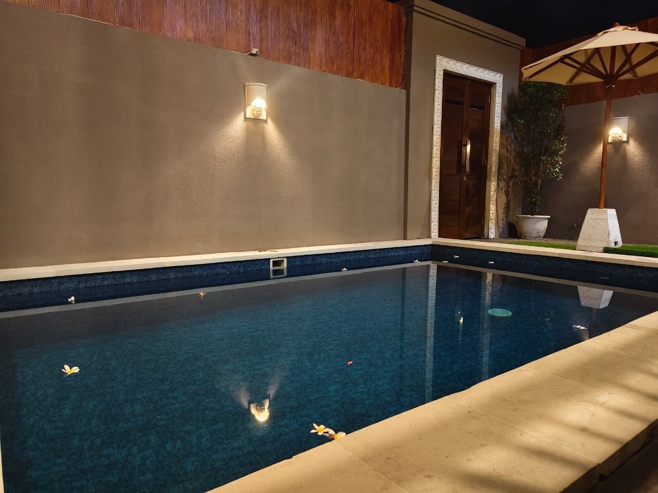 A swimming pool with a clear blue water surface, surrounded by a beige poolside area and a wooden wall.