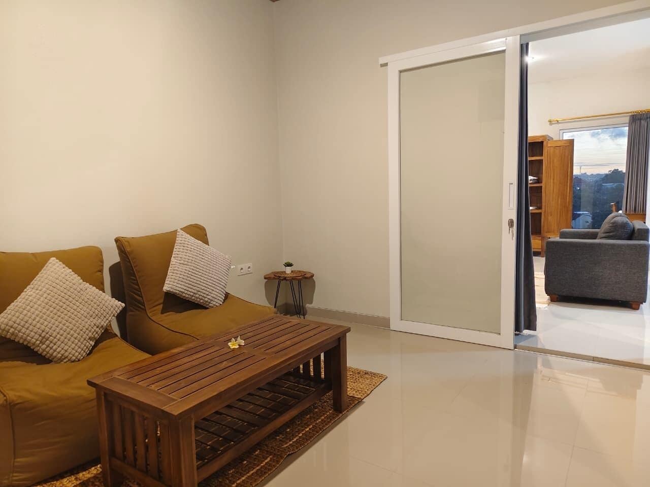 Una sala moderna con un juego de sofá, una mesa de centro de madera y un amplio ventanal corredizo que da a una habitación contigua con un sofá gris.