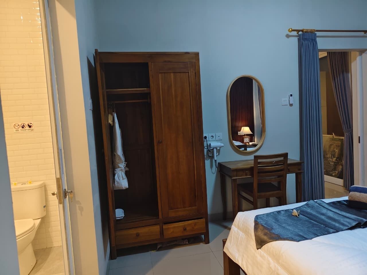 A bedroom with a bed, a wooden wardrobe, a desk with a chair, a mirror, and a bathroom with a toilet.