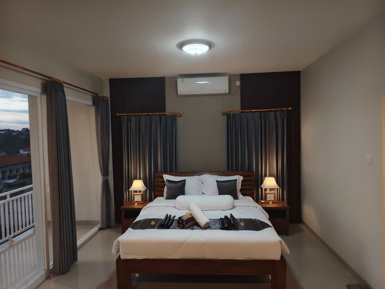 A hotel room with a bed, two bedside tables, a window with curtains, an air conditioning unit, and a view of buildings outside.