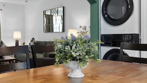 Un coin repas avec une table en bois, des chaises, un vase de fleurs, un ventilateur de plafond, un lave-linge fixé au mur et une œuvre d’art encadrée.