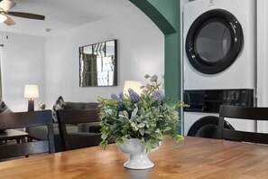 A dining area with a wooden table, chairs, a vase with flowers, a ceiling fan, a wall-mounted washing machine, and a framed artwork.