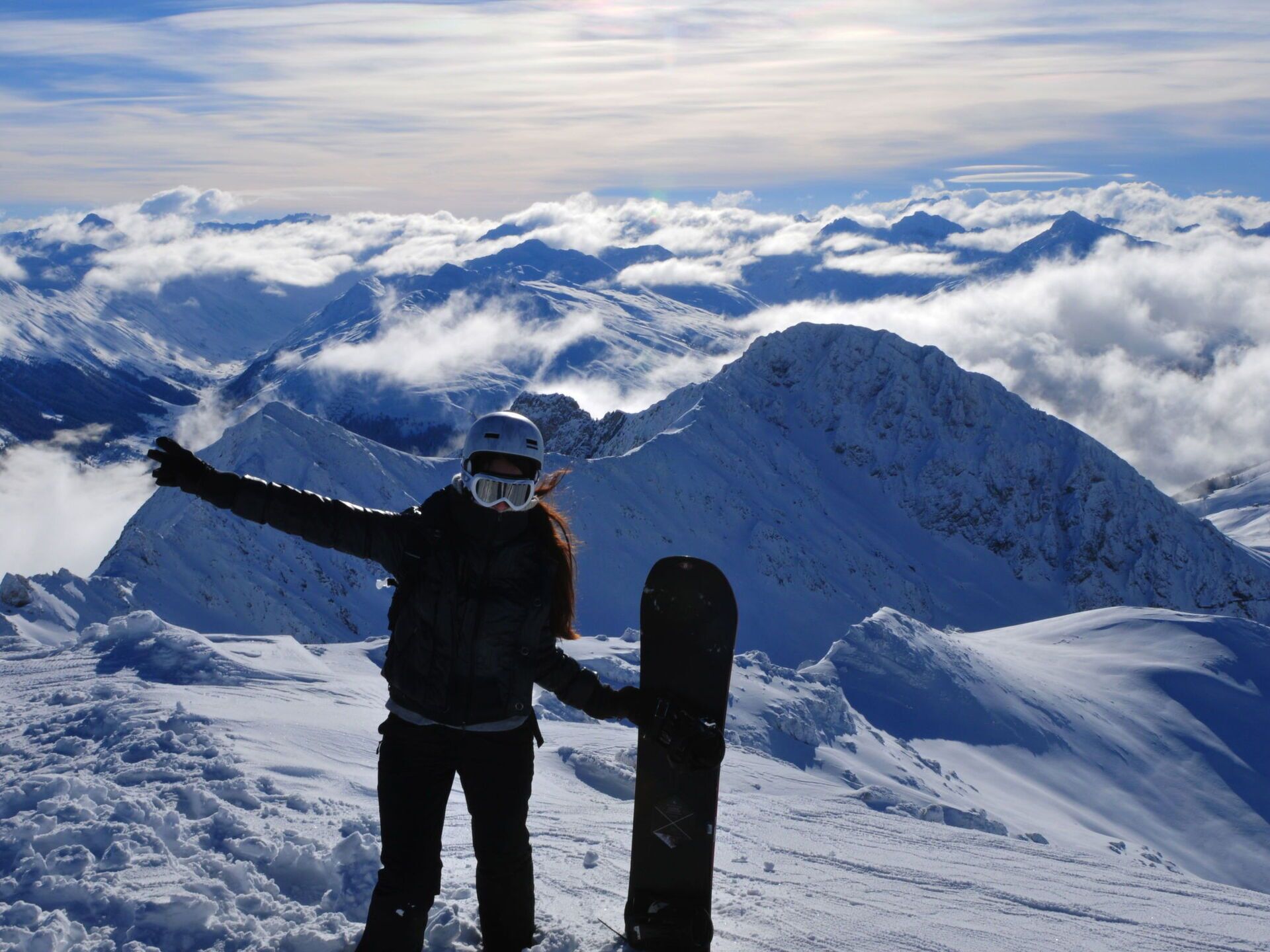 Eine Person steht mit einem Snowboard auf einem verschneiten Berg und blickt auf eine Bergkette mit Schnee und Wolken.