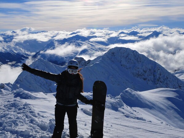 Een persoon staat op een besneeuwde berg met een snowboard, met uitzicht op een reeks met sneeuw bedekte toppen en wolken.