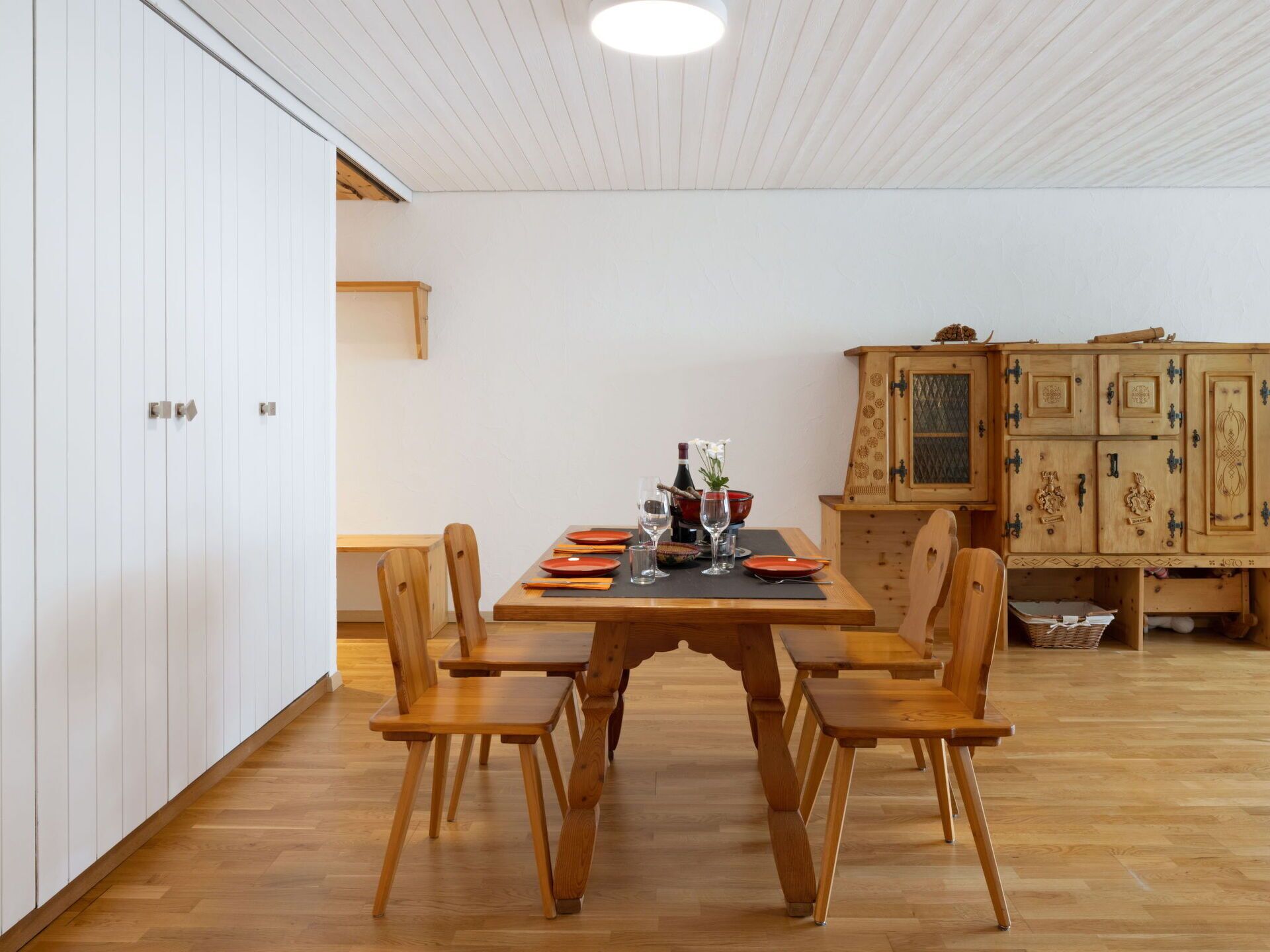 Ein Essbereich mit einem Holztisch, gedeckt für vier Personen, umgeben von Holzstühlen. Im Hintergrund ist ein Holzkasten mit aufwendigen Verzierungen zu sehen.