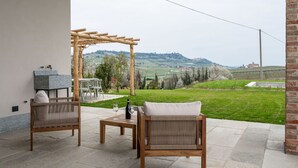 Un patio exterior con dos sillones de mimbre, una mesita y una botella de vino. Se distingue una pérgola al fondo, con vistas a un paisaje pintoresco con colinas y edificios.