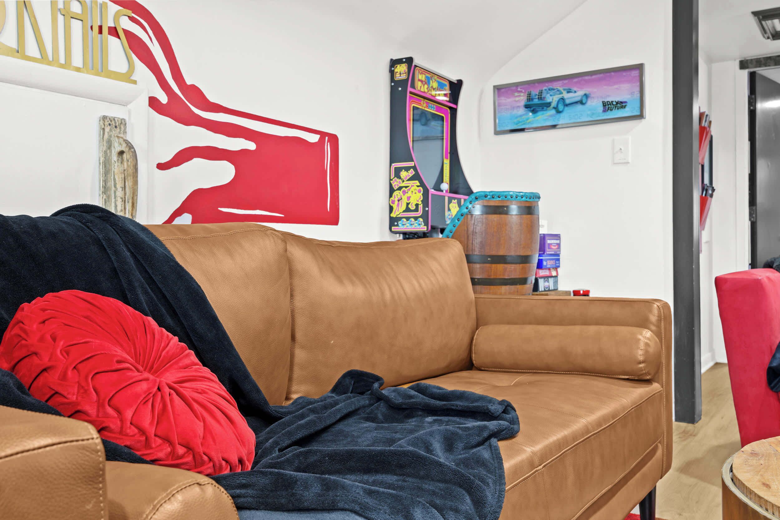 A room with a brown leather sofa, a red pillow, a blue blanket, a wooden barrel, a mirror, and a wall with a red abstract design and a framed picture.