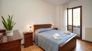 Un dormitorio con una cama de madera, una mesita de noche con lámpara, una planta en maceta y una ventana con cortinas.