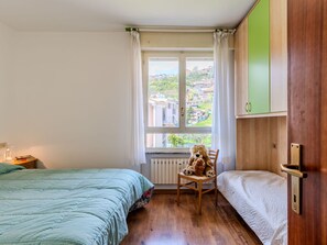 Une chambre avec deux lits, une chaise sur laquelle sont posés des peluches, une fenêtre donnant sur des immeubles et un radiateur.