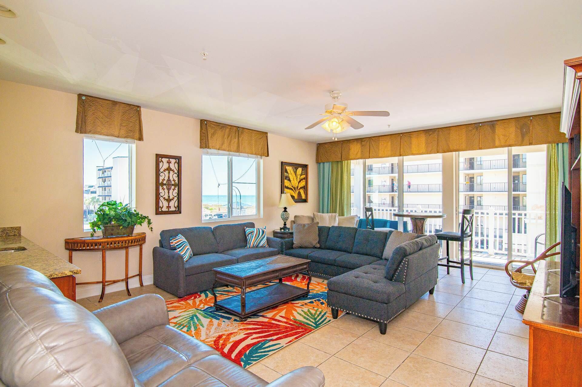 A spacious living room with a sectional sofa, a leather couch, and a coffee table. There are large windows with curtains, a ceiling fan, and a balcony with a table and chairs.