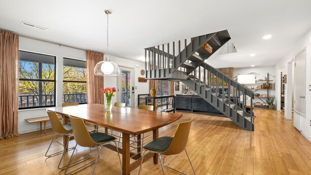 Un amplio comedor con una mesa y sillas de madera, una escalera moderna y ventanas grandes con cortinas.