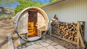 Una sauna de madera con puerta circular, un banco en su interior y un montón de leña afuera.
