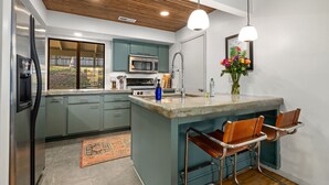 Una cocina moderna con gabinetes verde azulado, una isla central, electrodomésticos de acero inoxidable y techo de madera.