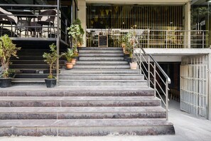Una escalera conduce a la entrada de un edificio, con mesas al aire libre y plantas en macetas.