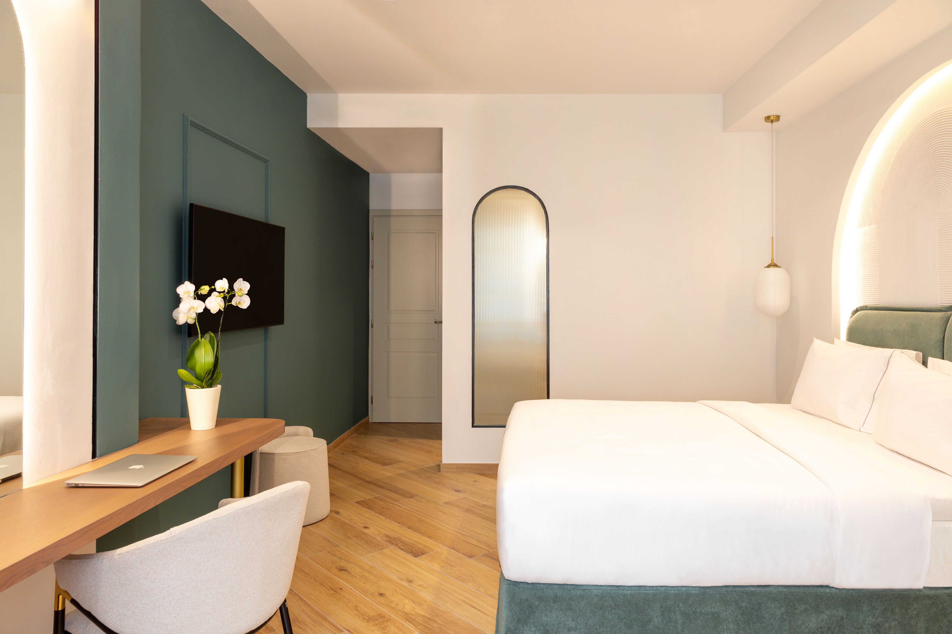 A modern hotel room with a wooden desk, a laptop, a white chair, a bed with white bedding, a green accent wall, a flat-screen TV mounted on the wall, and a small potted plant.