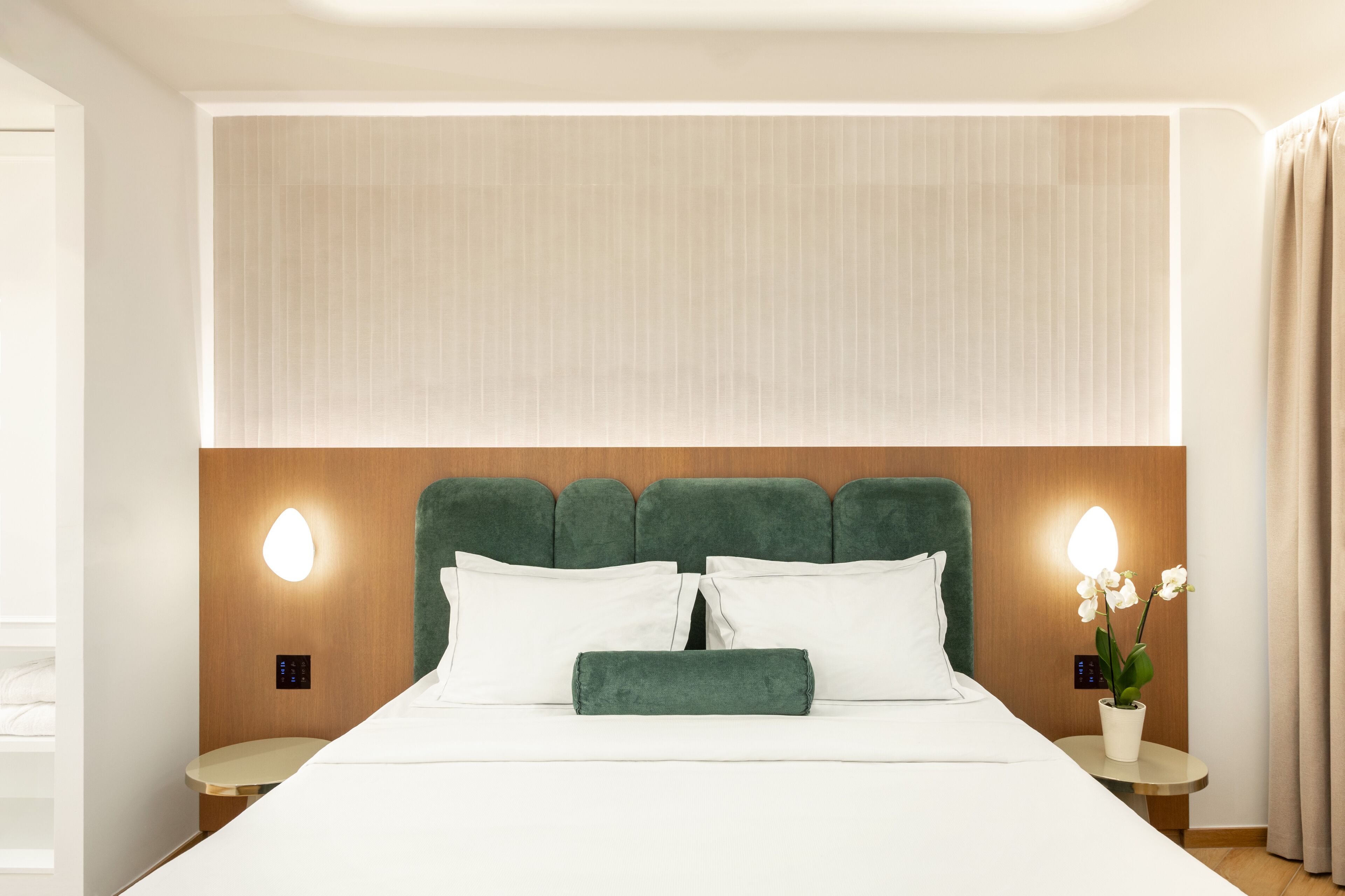 Cama bem arrumada com lençóis brancos, cabeceira estofada em verde e duas mesas de cabeceira com abajures.
