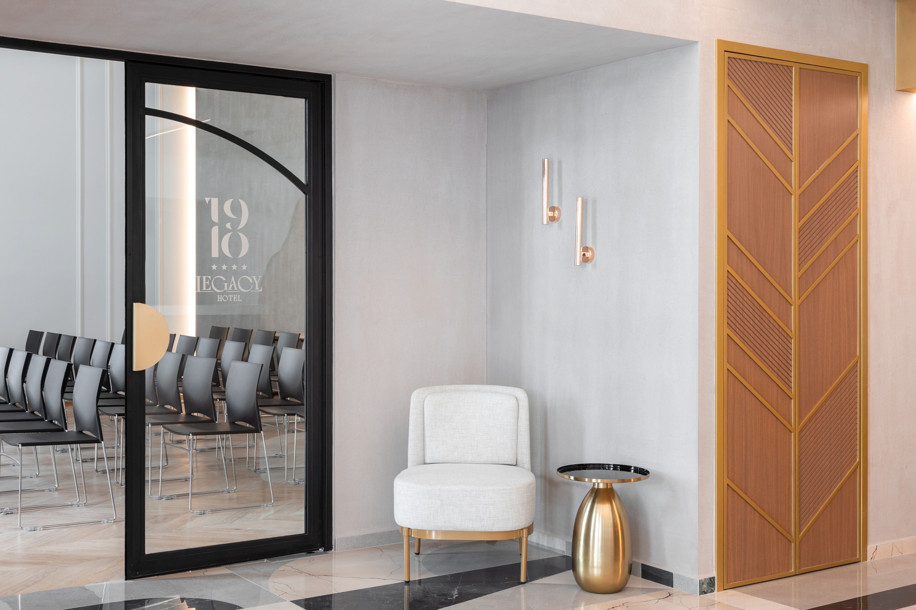 A modern hotel lobby with a seating area, a white armchair, a gold table, and a wooden door with a geometric design.