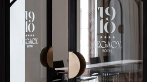 A hotel entrance with a glass door reflecting the interior. The door has a circular design element and the hotel name "LEGACY HOTEL" is visible on the glass. - 1910 Legacy Hotel (Volos)