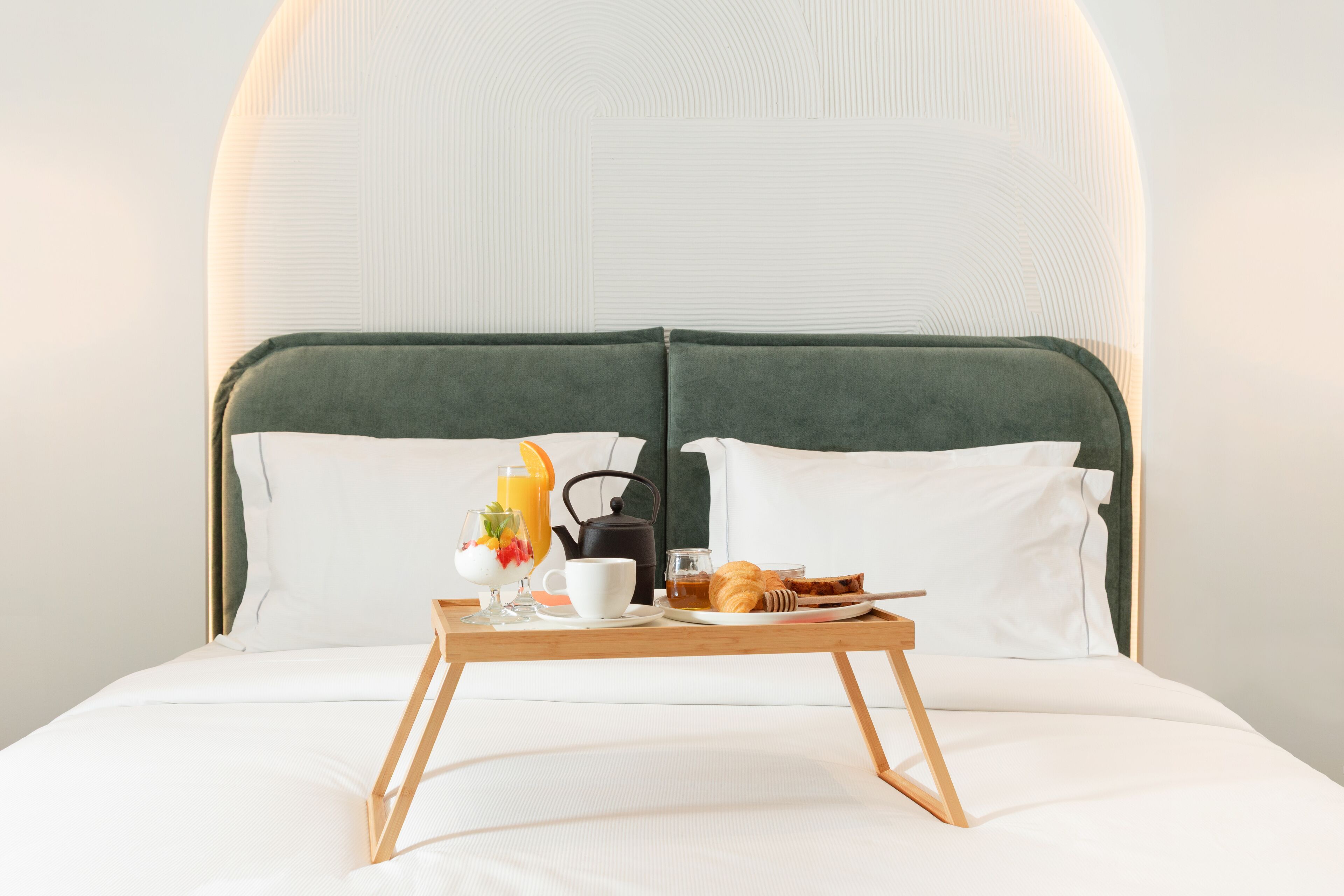 Un lit bien fait, avec un plateau contenant divers éléments pour le petit-déjeuner, notamment des croissants, une théière et un verre de jus d’orange.