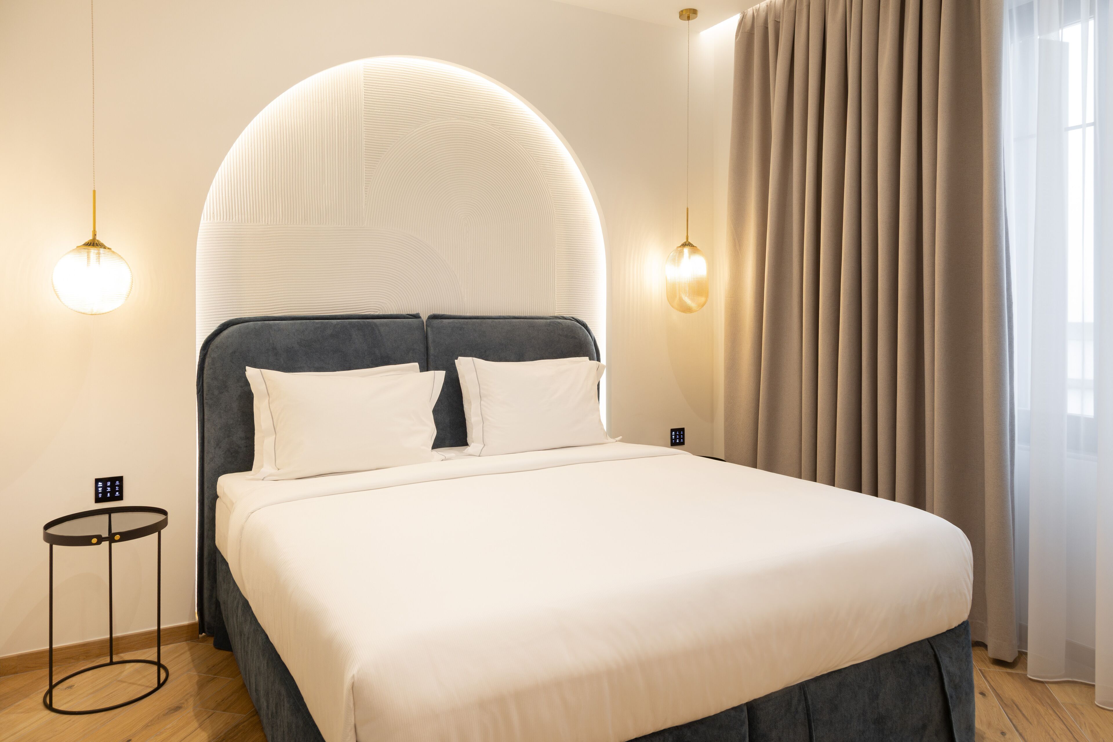 Una habitación de hotel moderna con una cama grande, un cabecero redondo, dos mesitas de noche y vistas al exterior a través de cortinas translúcidas.