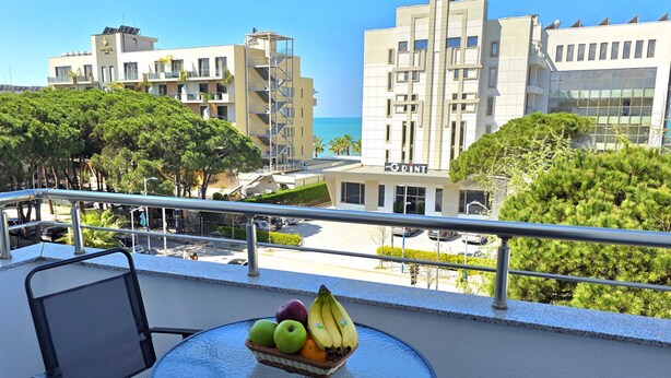 Een balkon met een tafel waarop een fruitmand staat, met uitzicht op een hotel en een helderblauwe lucht.