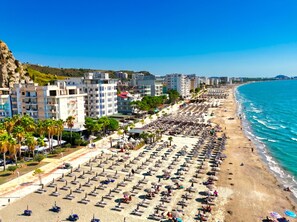 A beach with rows of sun loungers and umbrellas, buildings in the background, and a clear blue sky.