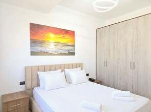 A bedroom with a bed, a nightstand, a wardrobe, and a painting of a sunset over the ocean. - Sunny Stay Apartment Durres 080 (Durres)