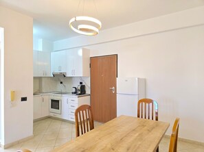 A compact kitchen with white cabinets, a wooden door, a built-in oven, a refrigerator, and a dining table with two chairs.