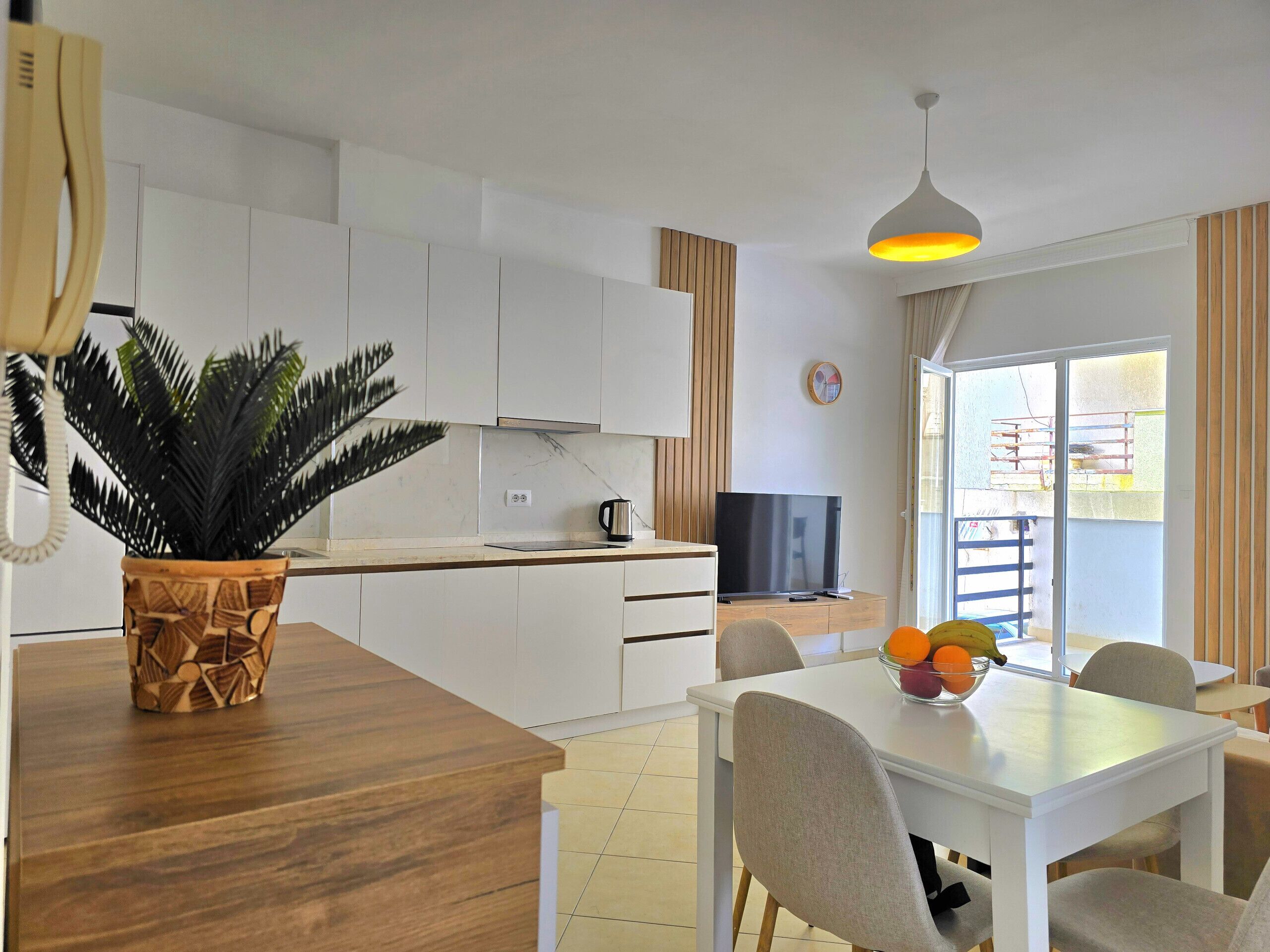 Une cuisine moderne avec des meubles blancs, un îlot central en bois, une plante en pot, une bouilloire et un téléviseur.