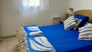 A bedroom with a blue bedspread, striped pillows, a wooden headboard, a bedside table, and a window with a white curtain. - Cozy house for 6, terrace, residence with pool and parking near the beach (Ondres)