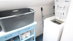 A laundry room with a white washing machine, a blue laundry cart with a basket, and a shelf with towels.
