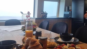 Une table de petit-déjeuner avec des croissants, des fruits et des boissons, un vase rempli de fleurs et une vue sur la mer par de grandes fenêtres.