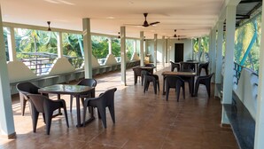 A spacious indoor area with multiple tables and chairs, large windows with a view of greenery, and ceiling fans. - Anandvan (Nagaon)
