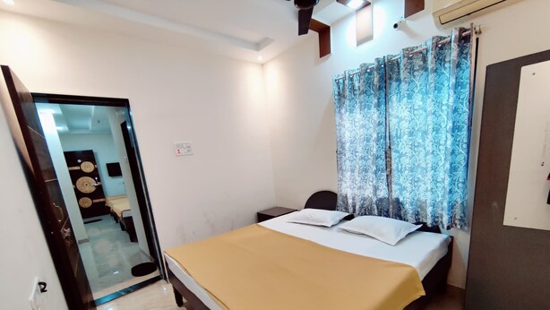 A bedroom with a bed, a nightstand, a mirror, and a door leading to a bathroom. - Anandvan (Nagaon)