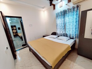A bedroom with a bed, a nightstand, a mirror, and a door leading to a bathroom. - Anandvan (Nagaon)