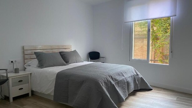 Ein Schlafzimmer mit einem Holzkopfteil, einem Bett mit grauer Bettwäsche, einem Nachttisch mit Schubladen, einem Stuhl und einem Fenster mit Blick ins Grüne.