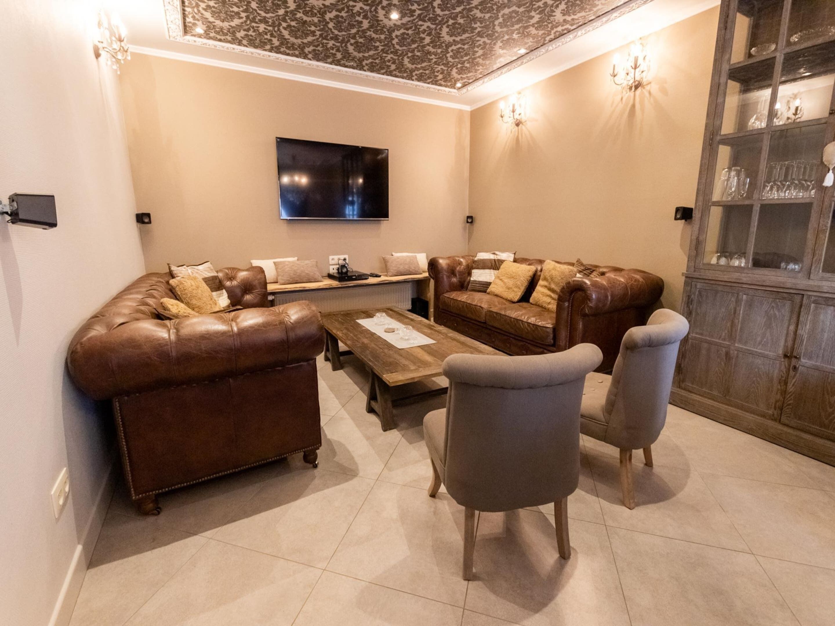 A living room with a brown leather sofa, a wooden coffee table, and two grey armchairs. A flat-screen TV is mounted on the wall above the sofa.
