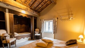 A cozy bedroom with a wooden ceiling, a large bed with a quilted cover, a chair, a window with curtains, and a lamp on a bedside table. - Villa in Coo With Indoor Pool & Near Waterfalls (Stavelot)