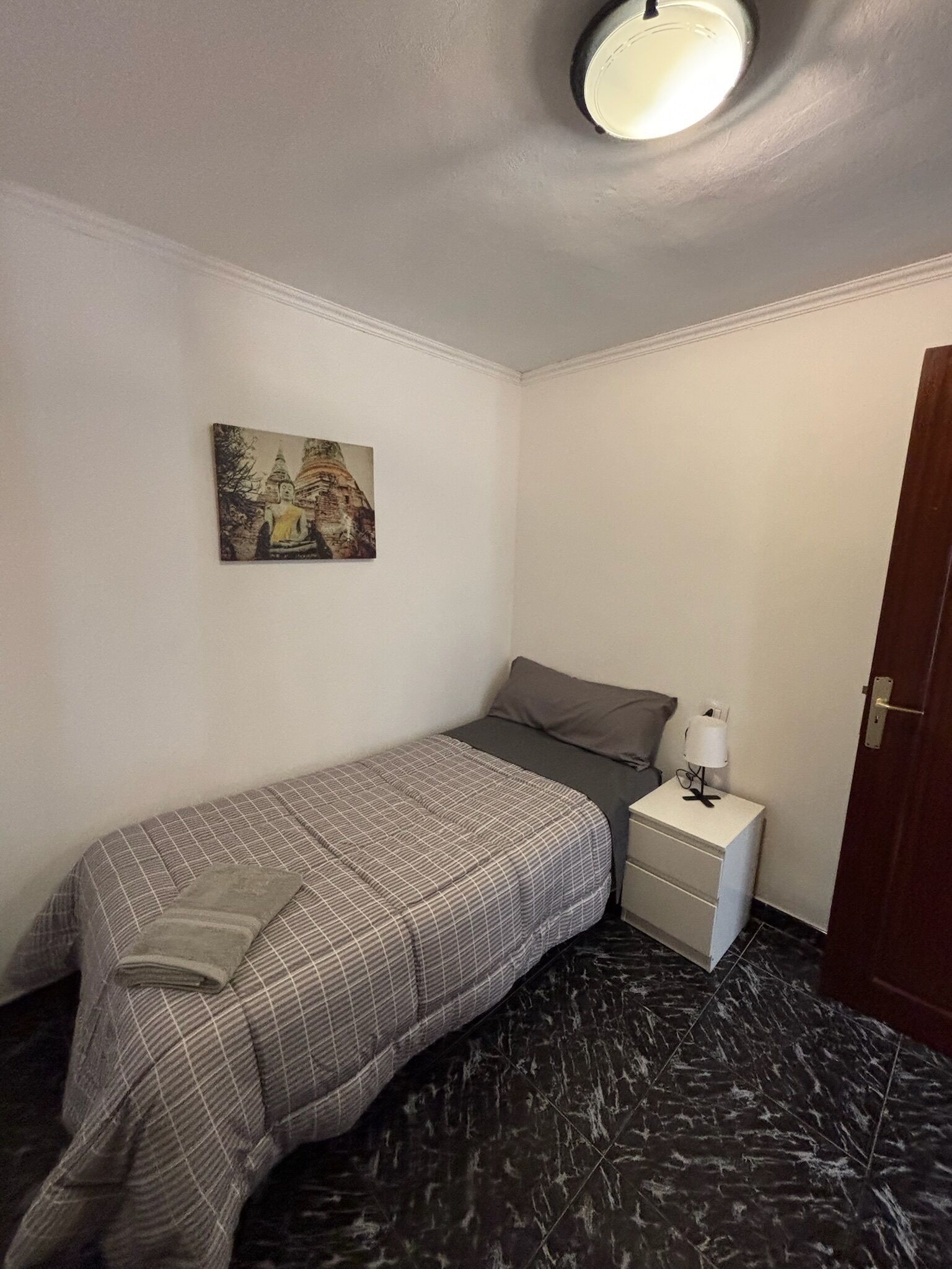 A small, neatly arranged bedroom with a bed, a nightstand, a lamp, and a painting on the wall.