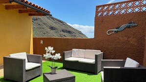 Un espace extérieur aménagé en terrasse, avec des meubles en osier, une petite table sur laquelle est posée une orchidée en pot, et un élément décoratif mural.