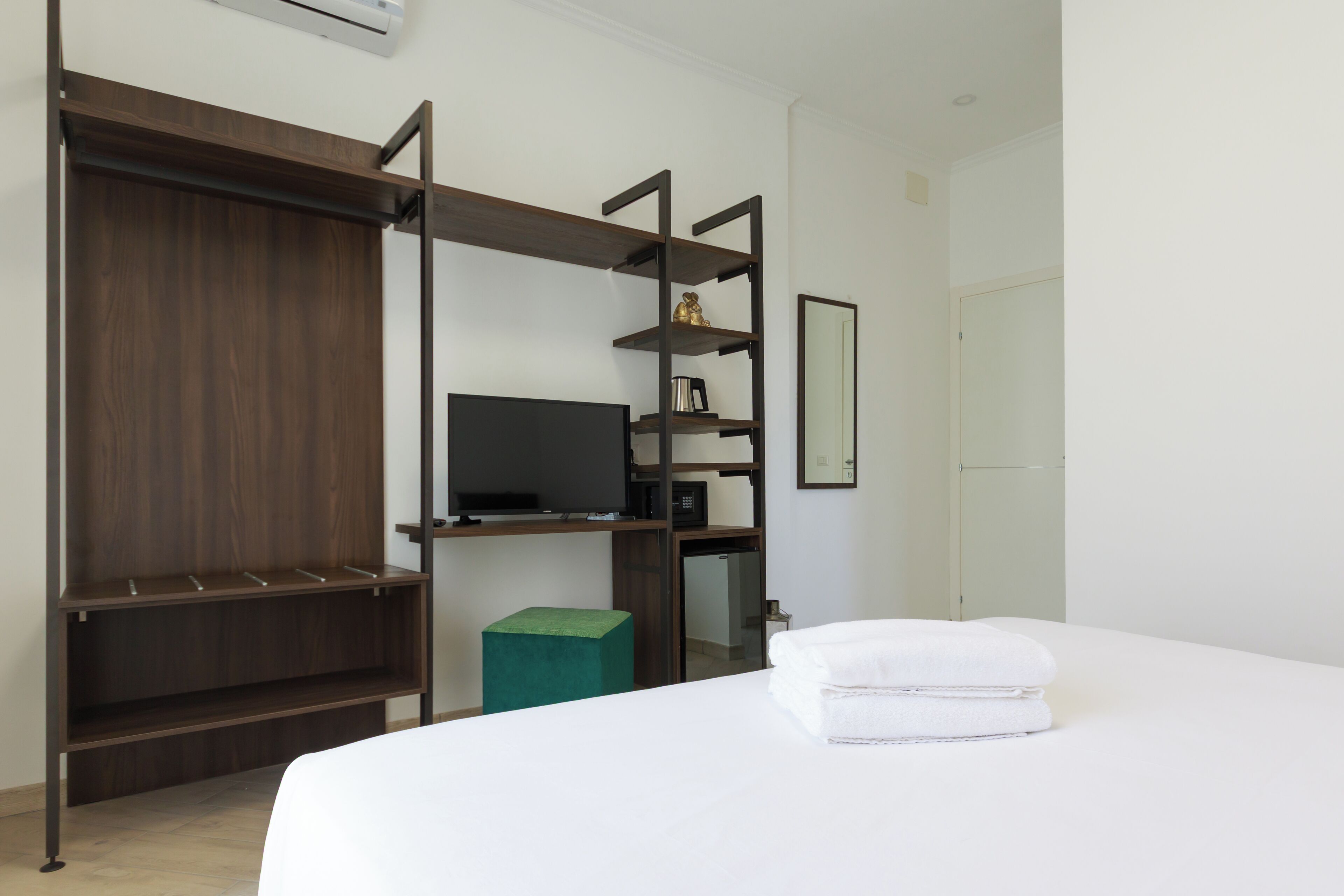 A modern hotel room with a large bed, a TV, a wardrobe, and a small green stool.