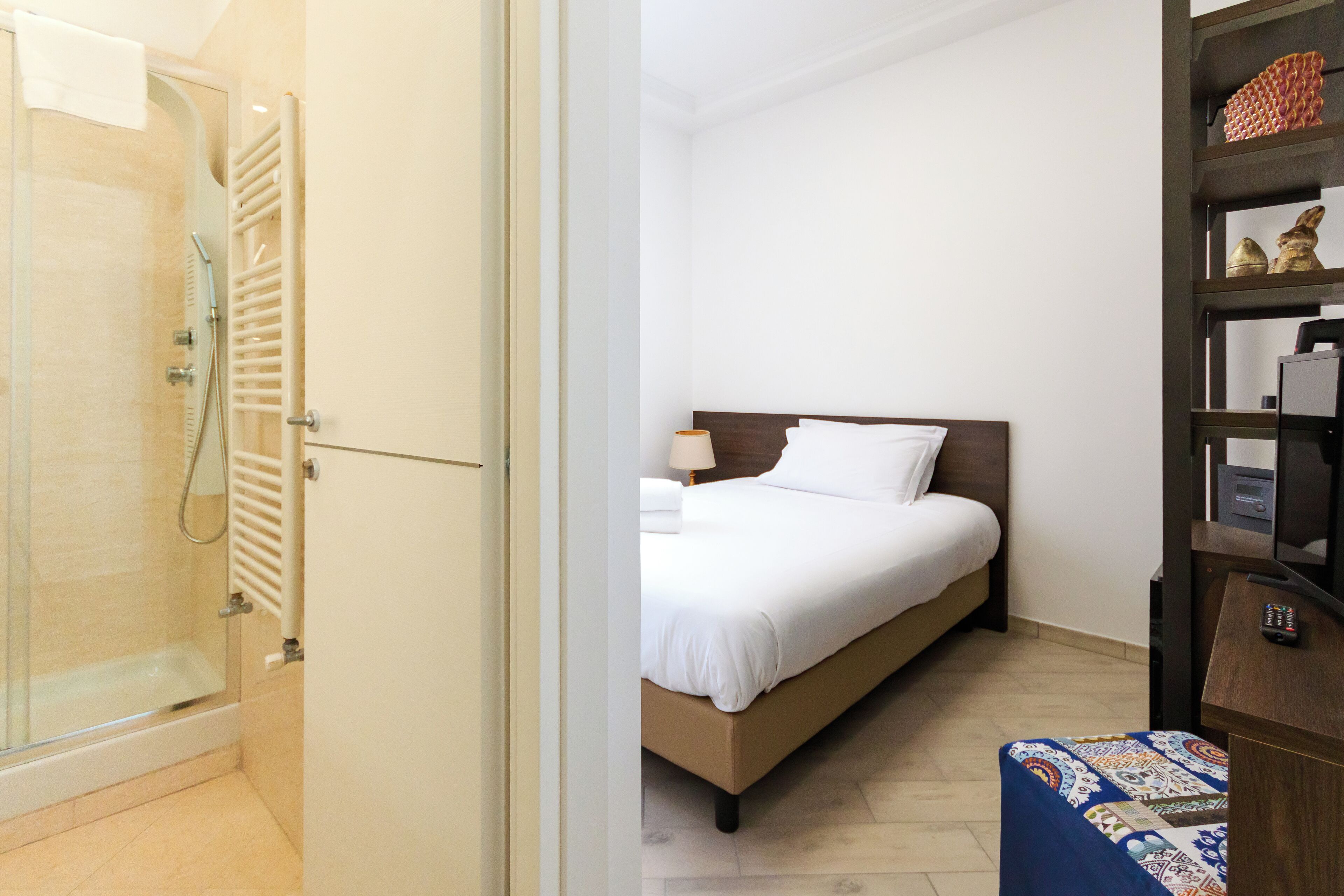 Camera d'albergo con doccia, letto con lenzuola bianche, comodino con lampada e un piccolo tavolino in legno con telecomando.