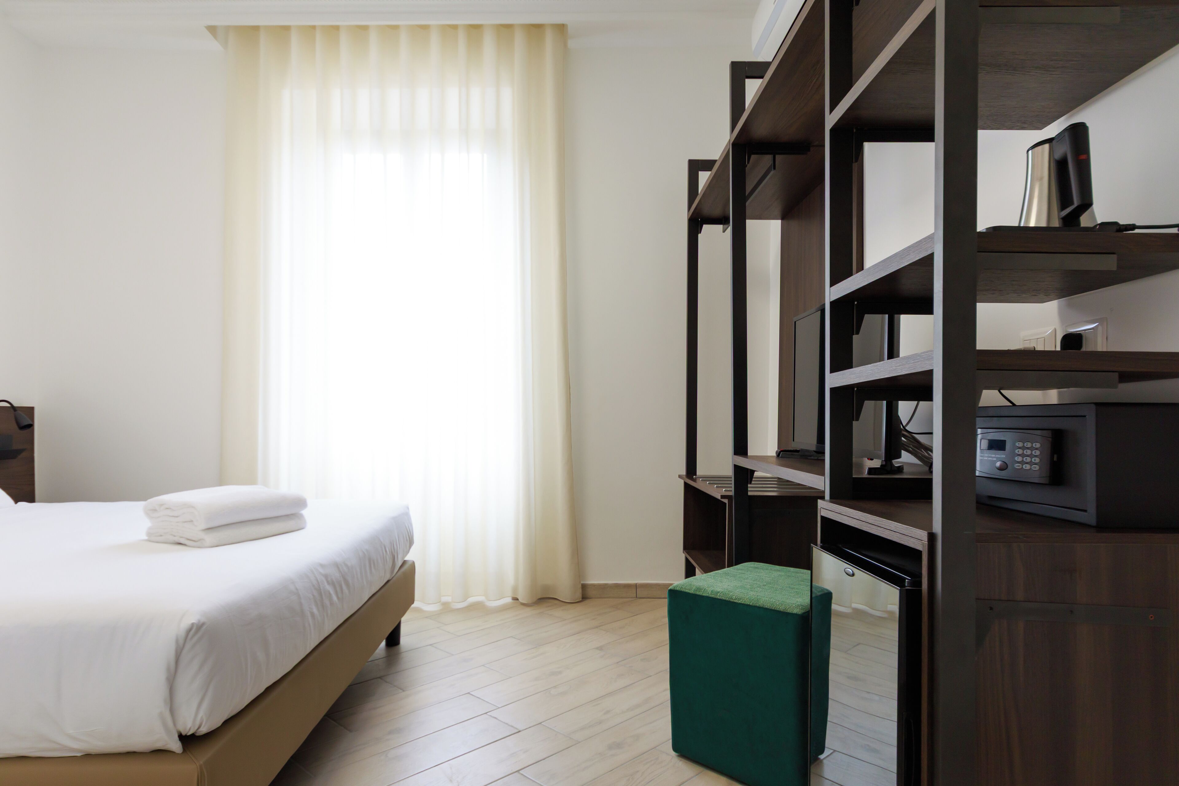 A hotel room with a bed, a green ottoman, a wooden bookshelf, and a microwave.