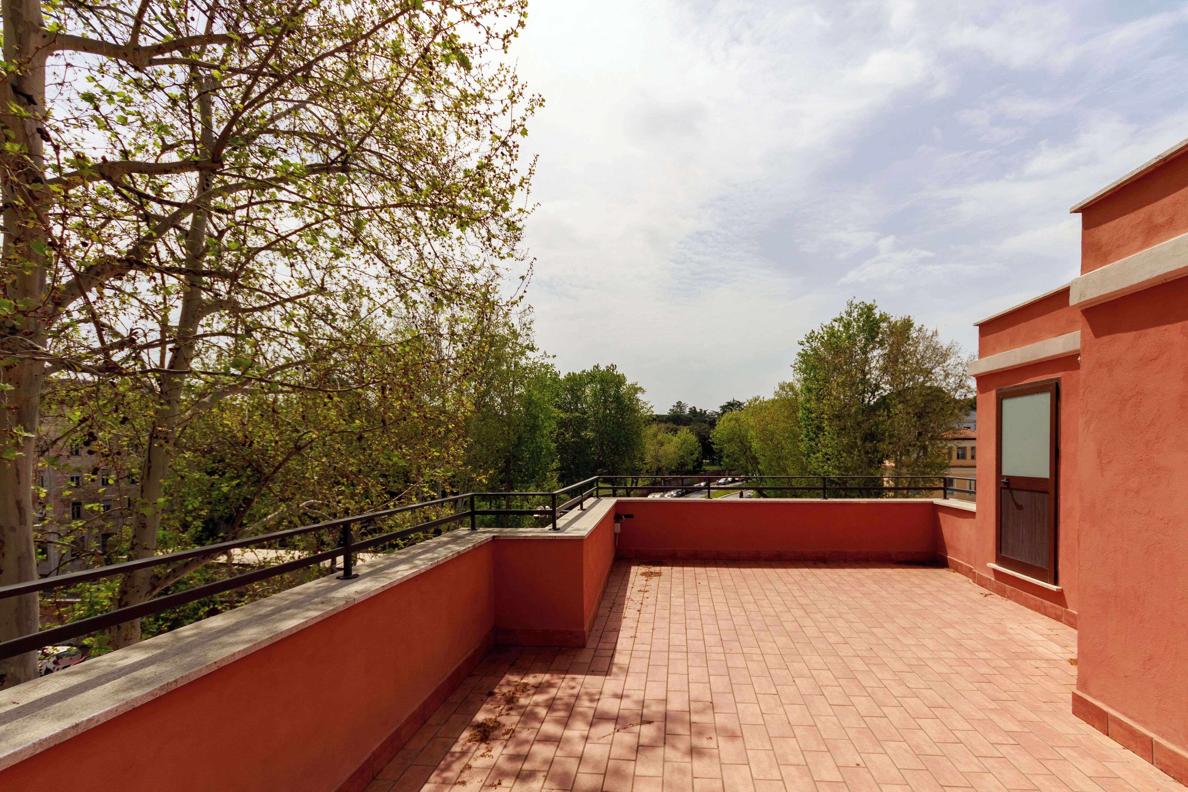 Terraço no telhado com chão de tijolos, rodeado por árvores e um edifício vermelho.