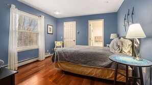 A bedroom with a bed, a round table, a lamp, a window with blinds, and a door.
