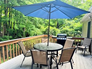 Een houten terras met een ronde tafel en stoelen, een barbecue en een blauwe parasol.