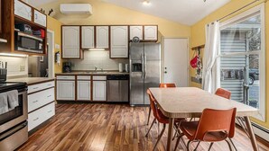A kitchen with wooden flooring, white cabinets, a stainless steel refrigerator, and a dining table with orange chairs.